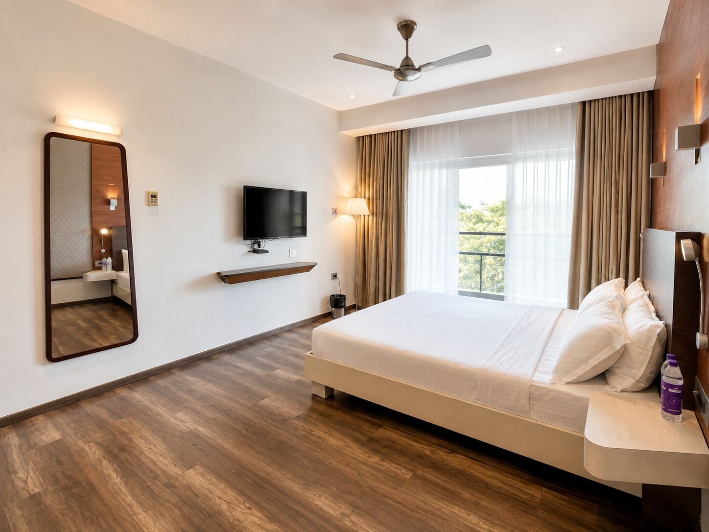 A hotel room with a bed, a television, a ceiling fan, a large window with curtains, and a wooden floor.