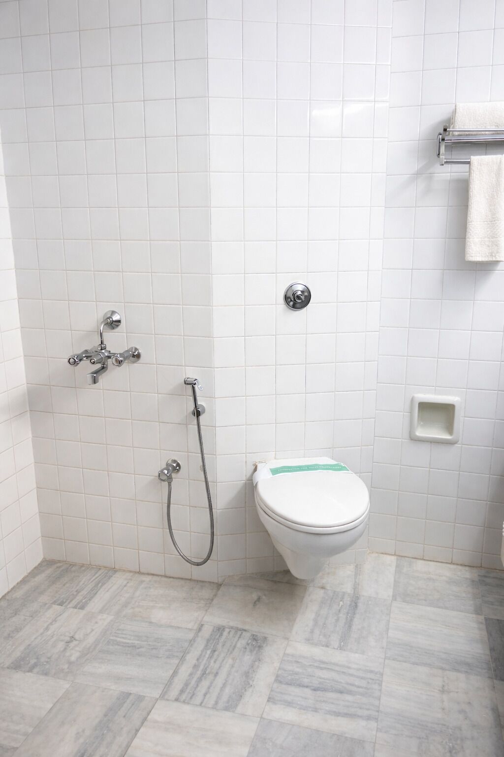 Un baño con paredes de azulejos blancos, un inodoro blanco, una ducha con manguera y un perchero para toallas.