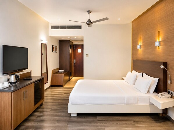 A modern hotel room with a large bed, a flat-screen TV, a desk with a coffee maker, and a ceiling fan.