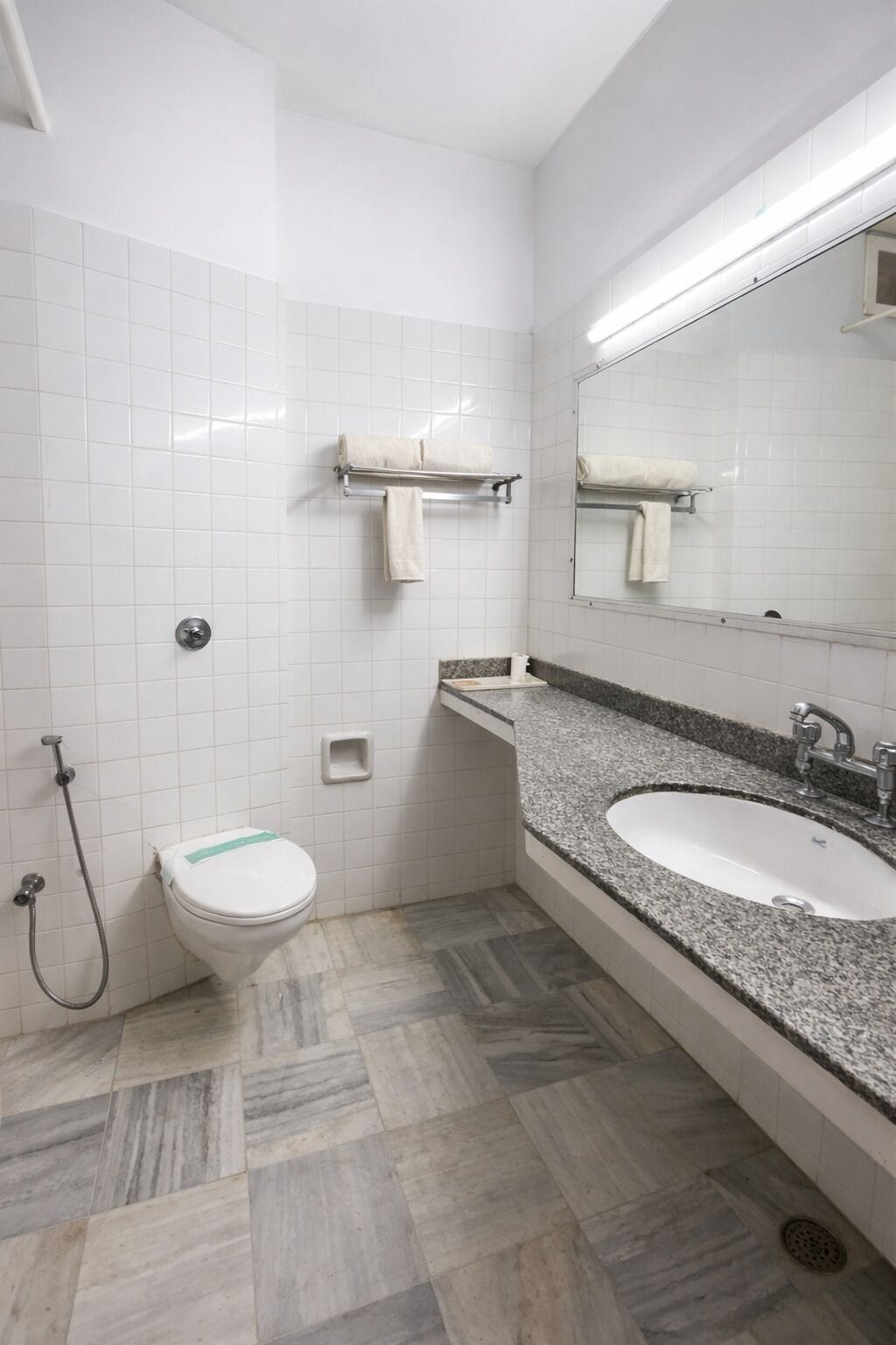 Un baño con paredes de azulejos blancos, un lavamanos de encimera de granito, inodoro, ducha y espejo.