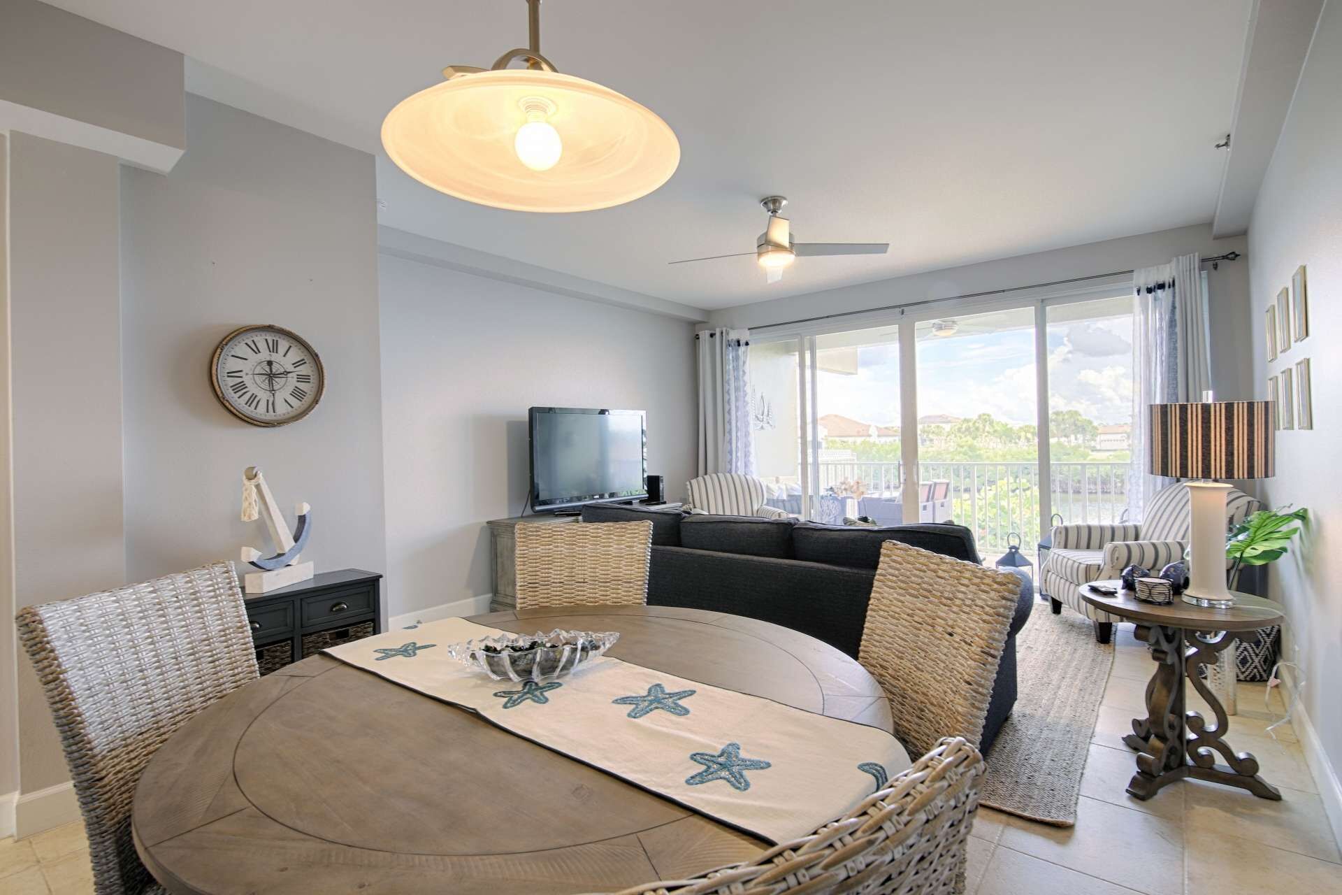 Un coin repas avec une table ronde, des chaises en osier, un ventilateur de plafond, un téléviseur et une vue sur l’extérieur grâce à de grandes fenêtres.