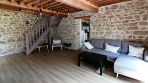 Ein rustikales Interieur mit einer Steinwand, Holzbalken, einer Treppe, einem grauen Sofa und einem schwarzen Couchtisch.