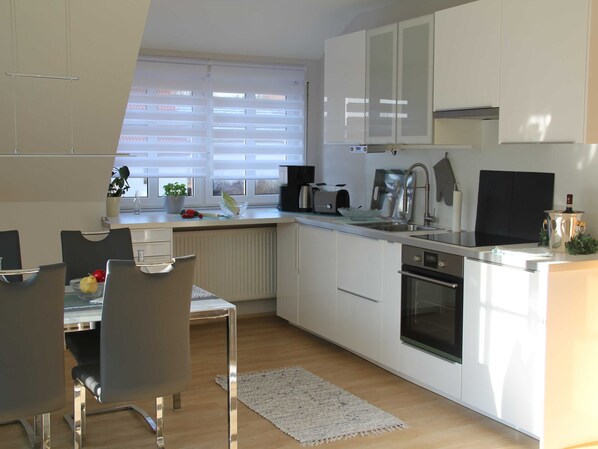 A modern kitchen with white cabinets, a dining table with chairs, a built-in oven, and a window with blinds.