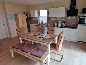 A modern kitchen with white cabinets, a built-in oven, and a dining area with a wooden table and cushioned chairs. - Ferienhaus Seglerblick am Achterwasser - Ferienhaus Seglerblick (Sauzin)