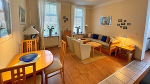Ein gemütliches Wohnzimmer mit Sofa, Sesseln, einem Esstisch und einem Fernseher. Fenster mit Vorhängen, eine Lampe und ein Bild an der Wand sind ebenfalls vorhanden.