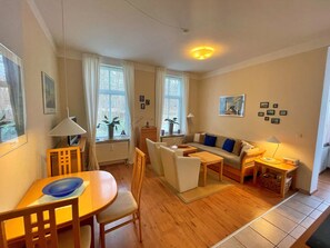 Ein gemütliches Wohnzimmer mit Sofa, Sesseln, einem Esstisch und einem Fernseher. Fenster mit Vorhängen, eine Lampe und ein Bild an der Wand sind ebenfalls vorhanden.