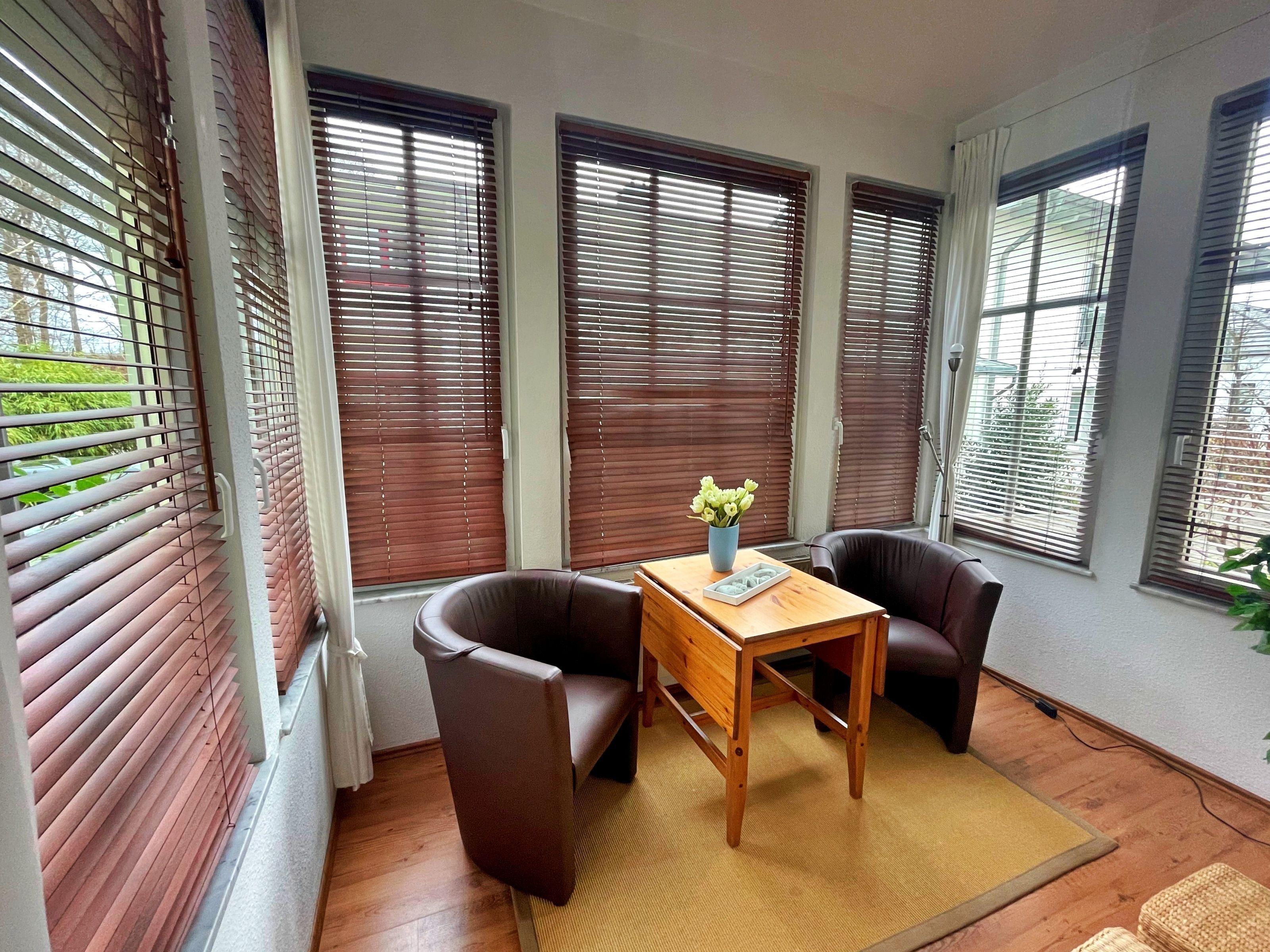 A room with a wooden table and two leather chairs, large windows with blinds, and a small plant on the table.