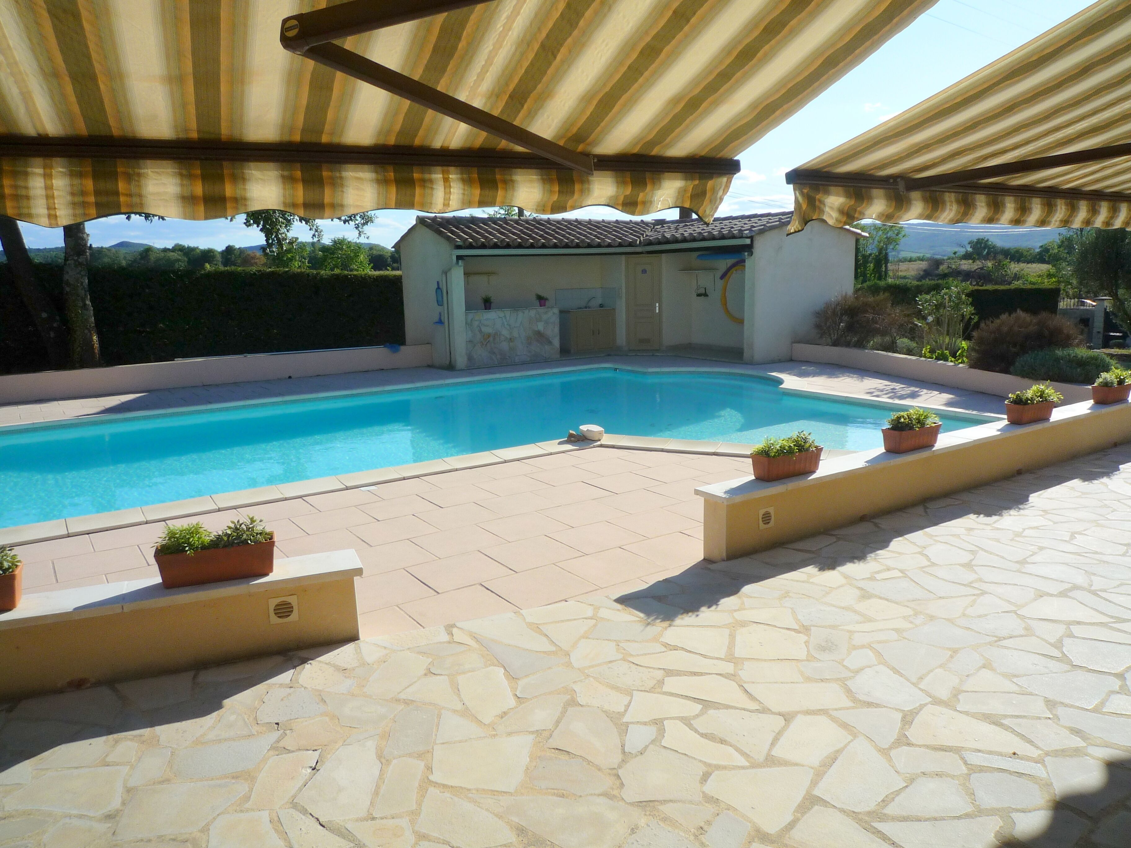 Una piscina con solarium in pietra, circondata da piante in vaso e un'area coperta con tende a righe.