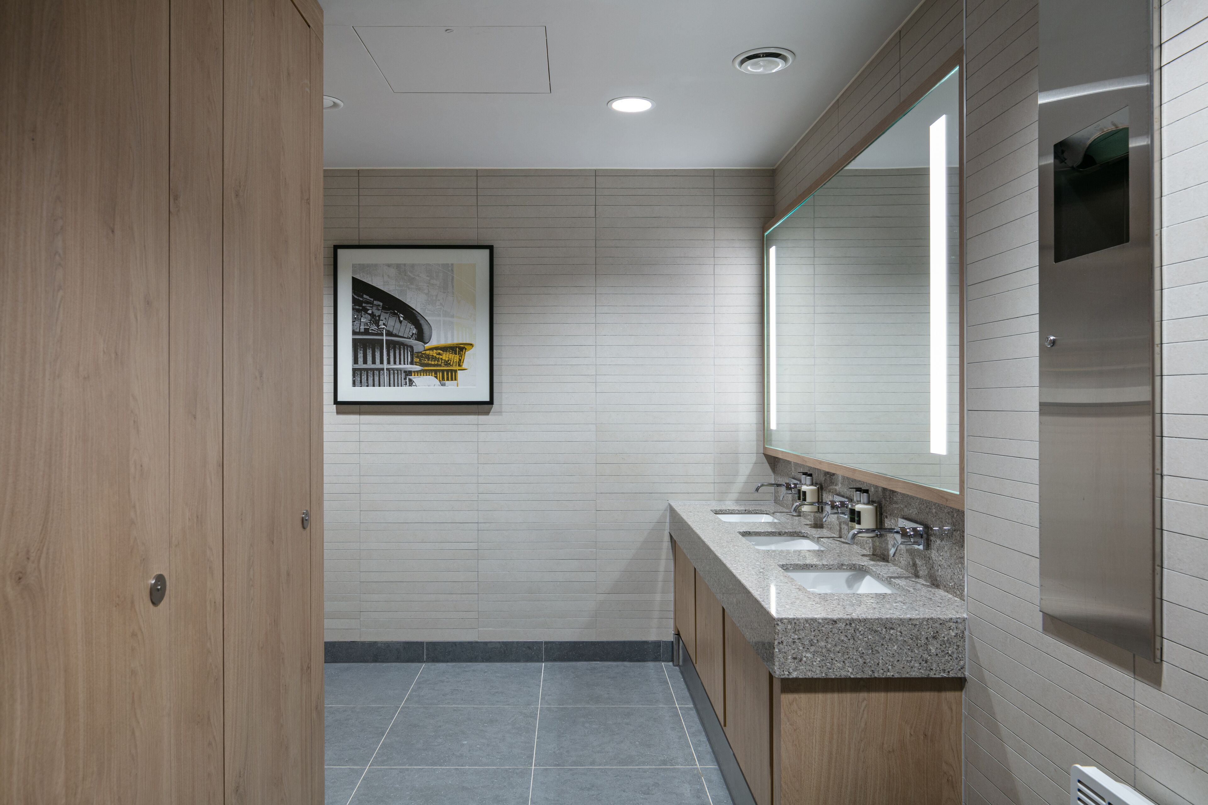 A modern bathroom with a wooden door, a framed picture on the wall, and a vanity with three sinks.