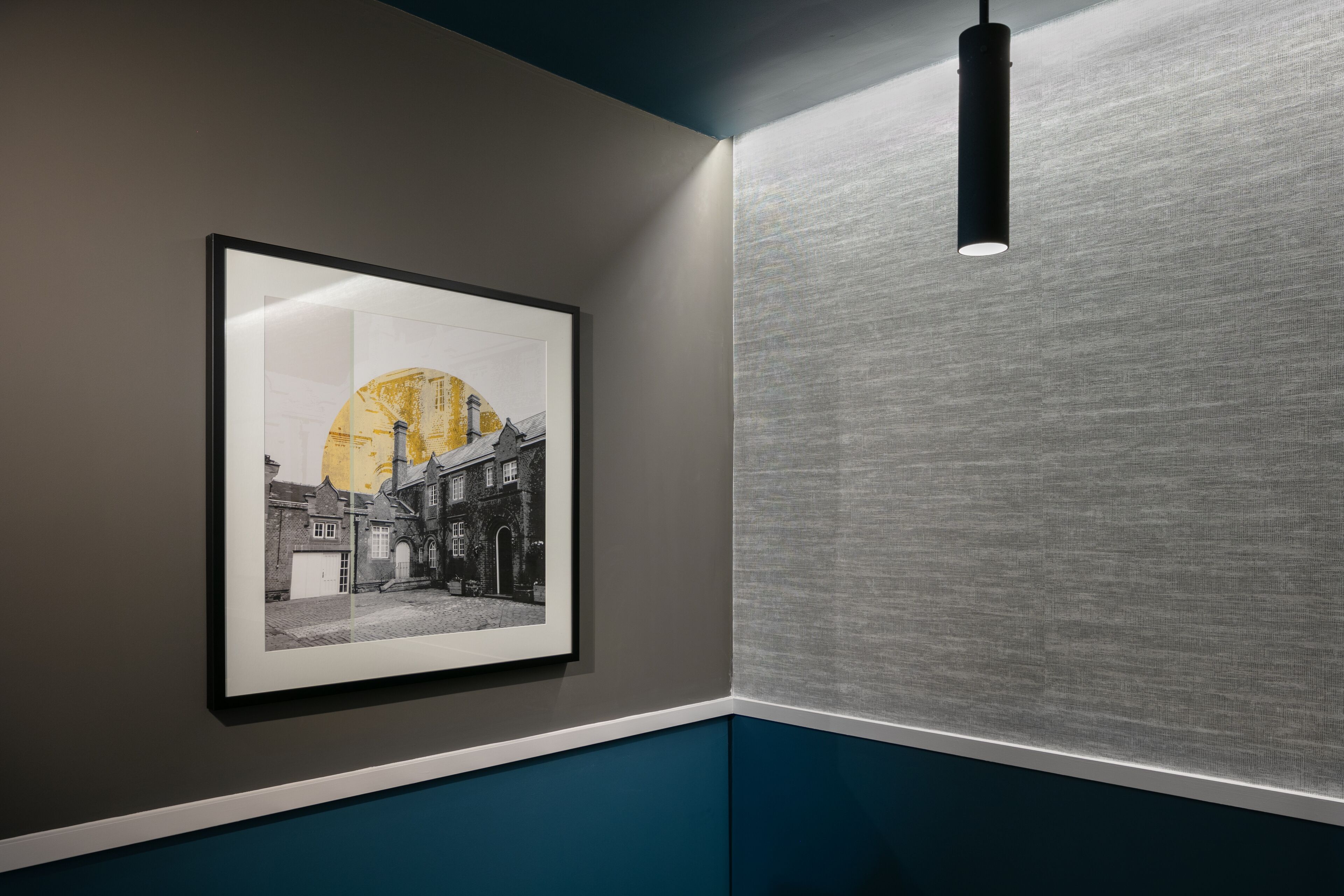 A framed black and white photograph of a street scene with a large yellow sun, mounted on a wall with a textured grey panel and a blue lower section.