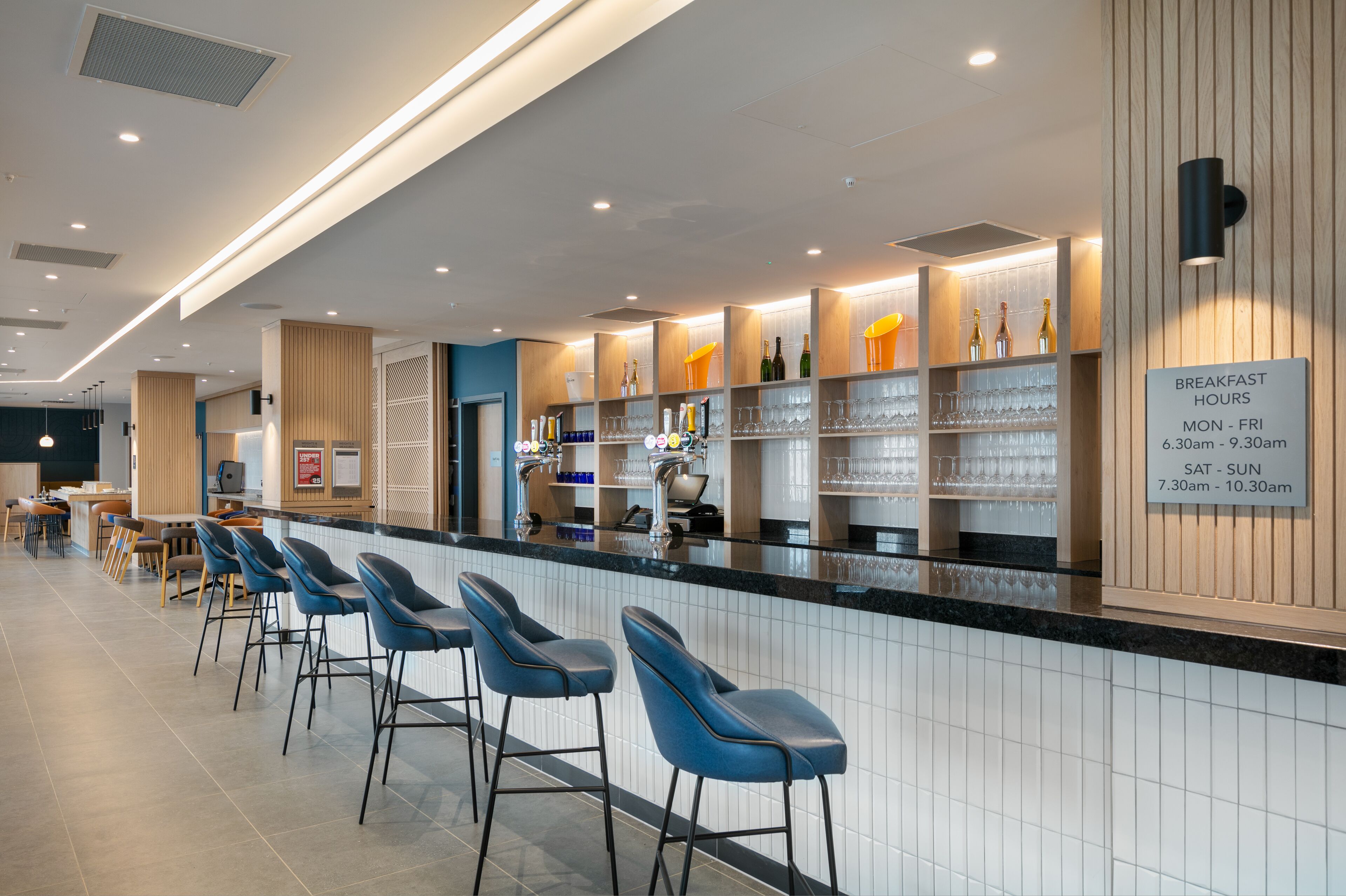 A modern bar area with a counter, bar stools, and shelves of bottles and glasses. A sign indicates breakfast hours.