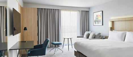 A hotel room with a large bed, a desk with a chair, a small table, and a TV.