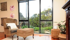 A room with a large window, a brown armchair, a matching ottoman, and two woven side tables with plants. - Castle on the Hill, Ocean Grove. (Ocean Grove)