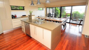 A modern kitchen with a central island, stainless steel appliances, and a large window overlooking a balcony. - Castle on the Hill, Ocean Grove. (Ocean Grove)