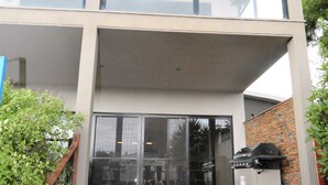 A modern two-story building with a covered outdoor area, a wooden staircase, and a barbecue grill. - Castle on the Hill, Ocean Grove. (Ocean Grove)