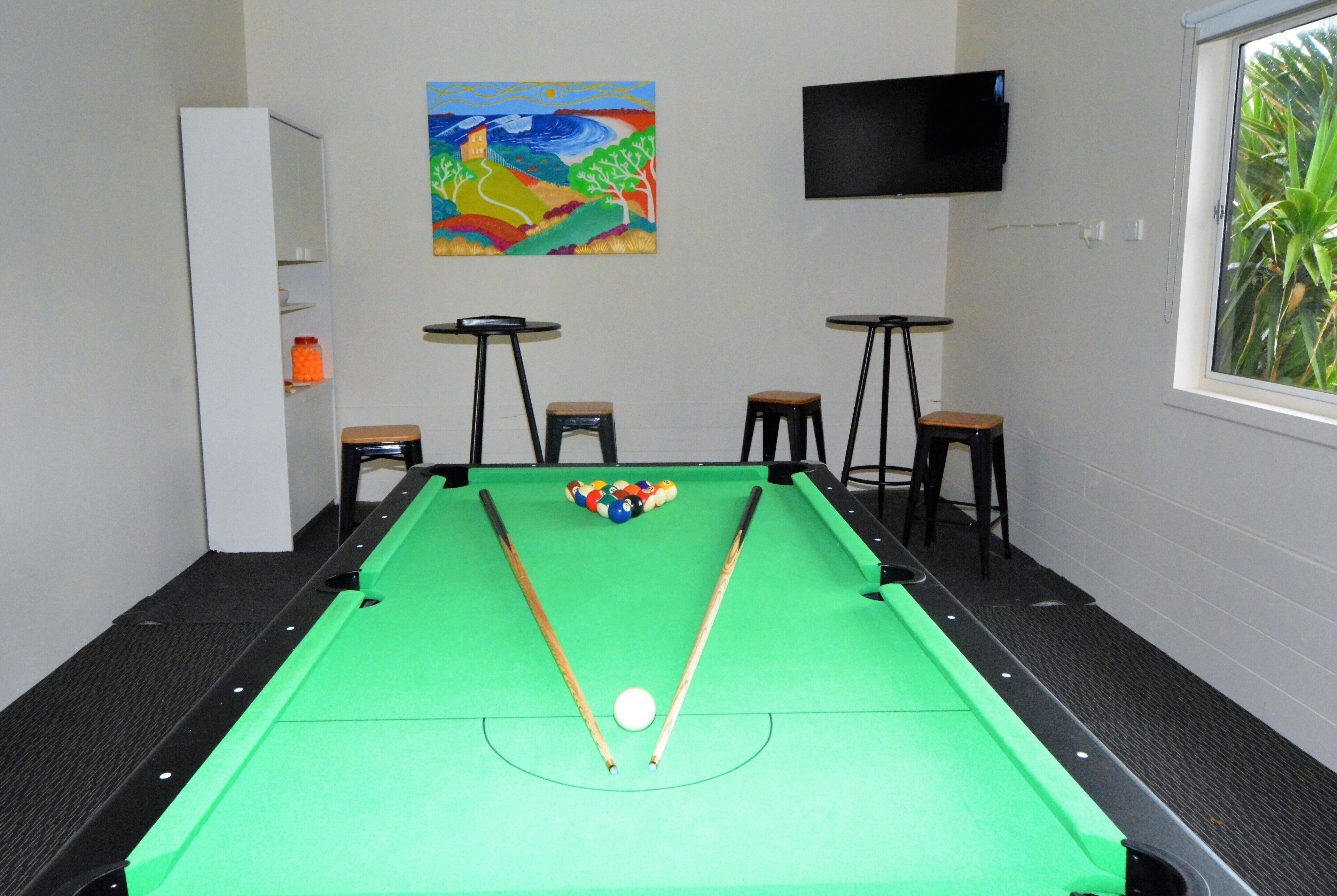 A pool table with a green felt surface, two pool cues, and a white cue ball. There is a colorful painting on the wall above the table and a television mounted on the wall to the right.