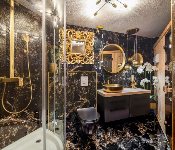 Un baño moderno con cabina de ducha de vidrio, tocador con lavabo dorado y espejo con marco decorativo.