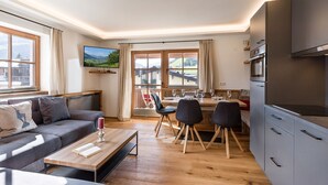 Ein modernes Wohnzimmer mit Sofa, Couchtisch und Essbereich mit Stühlen. Ein Fernseher ist an der Wand befestigt, und es gibt einen Blick auf einen Balkon mit Tisch und Stühlen.