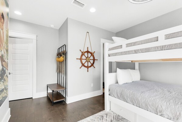 A room with a bunk bed, a wall-mounted ship's wheel, a small table with a plant, and a door with a painting.