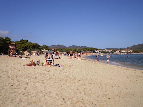 A beach with people sunbathing and swimming, a lifeguard tower, and houses in the background.