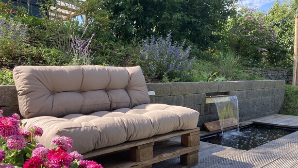 Une terrasse en bois avec un espace salon agrémenté de coussins et un petit élément décoratif aquatique.