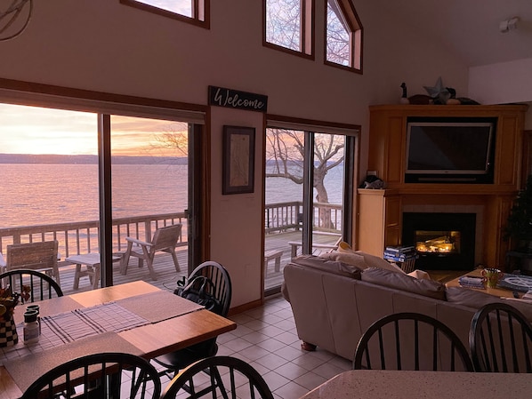 A cozy living room with a fireplace, a sofa, and a dining area with a table and chairs. There is a large window offering a view of the water and a deck outside.