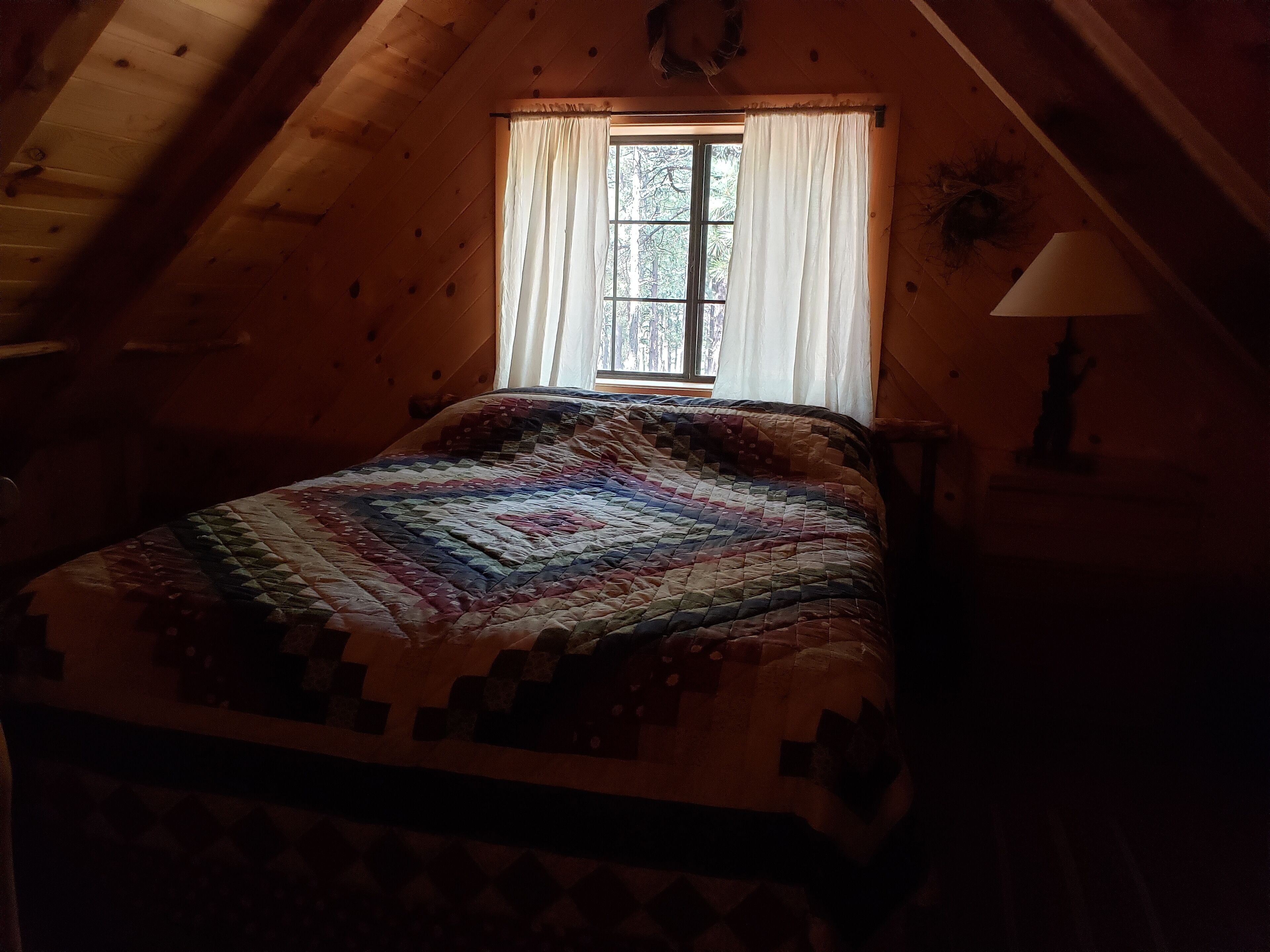 A cozy attic bedroom with a wooden sloped ceiling, a large window with white curtains, and a colorful quilt on the bed.