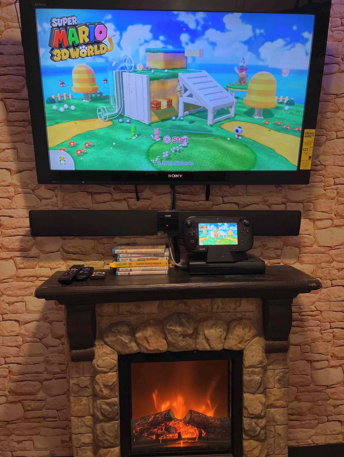 Kiviseinälle on asennettu televisio, jossa näkyy peli nimeltä ”Super Mario 3D World”. Sen alla, takan reunalla, on pelikonsoli ja pelilevy, ja takassa palaa tuli.