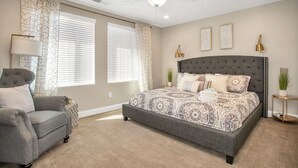 Ein Schlafzimmer mit einem großen Bett, einem grauen Sessel, einem Deckenventilator und einem Fenster mit Jalousien.