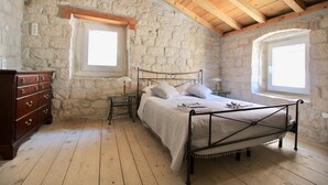 Ein Schlafzimmer mit Steinwand, Holzbalkendecke, einem Metallbettgestell, einer Holzkommode und zwei Nachttischen mit Lampen.