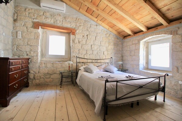 A bedroom with a stone wall, wooden ceiling beams, a metal bed frame, a wooden dresser, and two bedside tables with lamps.