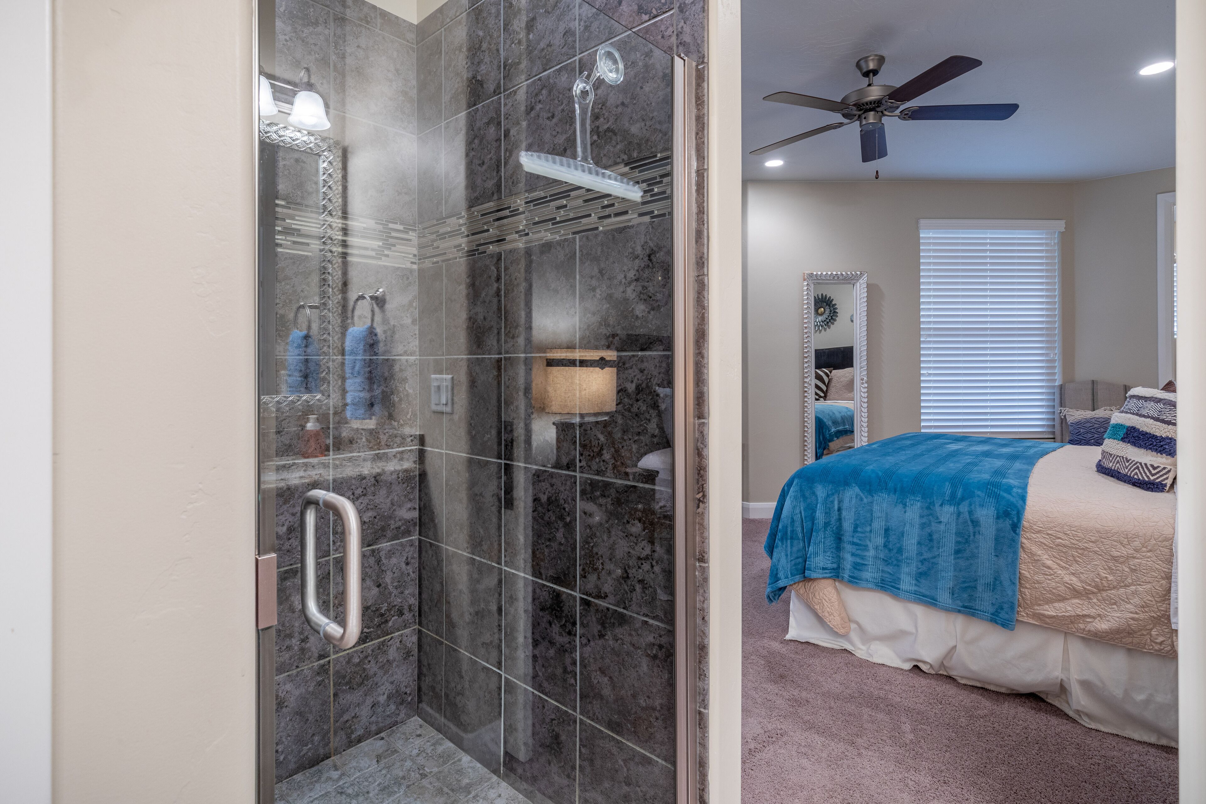 A modern bathroom with a glass-enclosed shower, a ceiling fan, and a bed with a blue blanket.