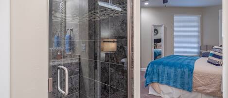 A modern bathroom with a glass-enclosed shower, a ceiling fan, and a bed with a blue blanket.