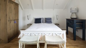 A bedroom with a wooden wardrobe, a bed with striped bedding, two wicker chairs, and a small table with a lamp. - Camber Sands House
Luxury 5 En-Suite bedrooms, minutes from beach. Dog friendly! (Camber)