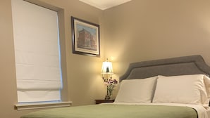 Ein ordentlich bezogenes Bett mit einer grünen Tagesdecke, ein Nachttisch mit Lampe und Blumen sowie ein gemustertes Teppich auf dem Boden.