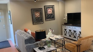 Ein Wohnzimmer mit grauem Sofa, einem Couchtisch mit Dekoration, einem Fernseher auf einem Schrank und gerahmten Bildern an der Wand.