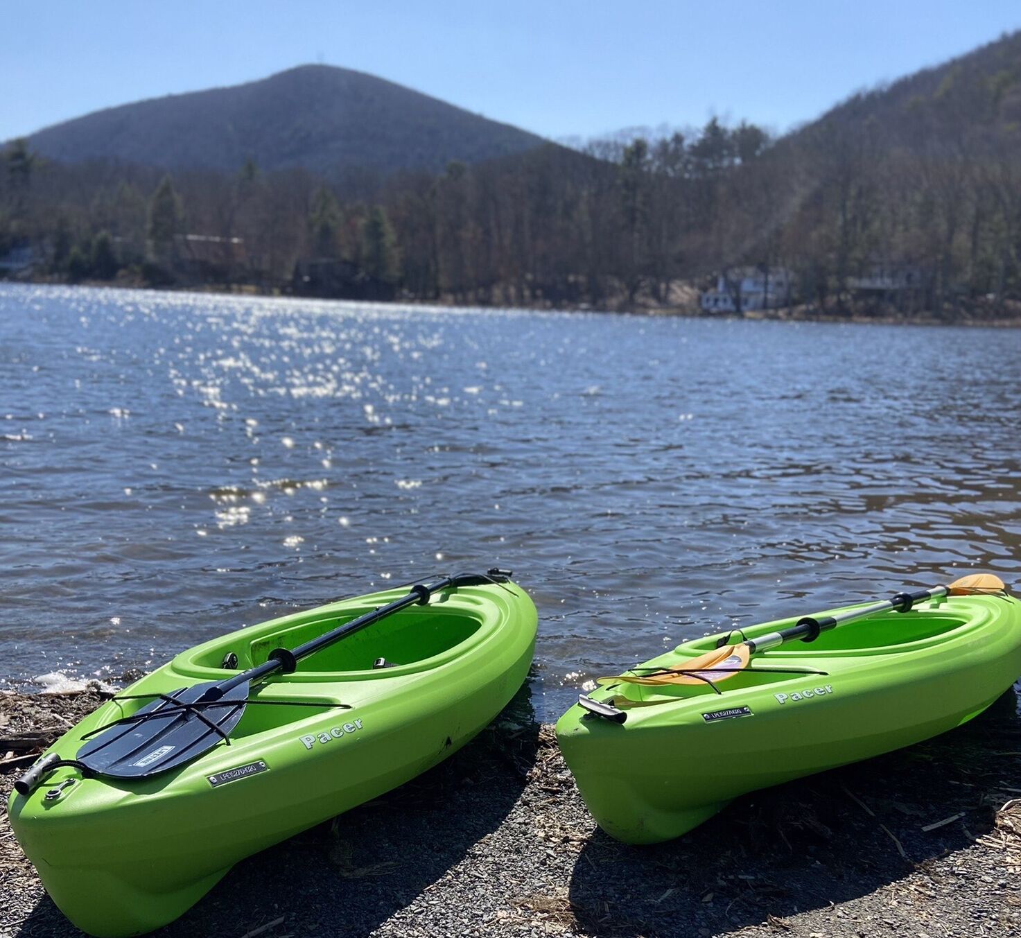 Two green kayaks with the brand 'Pacer' on a rocky shore by a lake with mountains in the background.