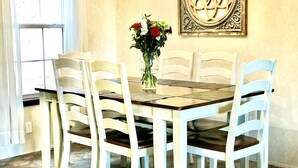 Ein Essbereich mit einem für sechs Personen gedeckten Tisch, umgeben von weißen Stühlen mit dunkel gefärbten Kissen. Auf dem Tisch steht eine Vase mit Blumen.