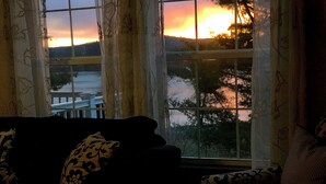 Ein Zimmer mit Sofa und Ohrensessel, zugewandt zu einem Fenster mit Blick auf einen Sonnenuntergang über Wasser und Hügeln.