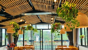 A spacious dining area with wooden tables and chairs, large windows offering a view of greenery outside, and hanging plants adding a touch of nature. - Rustic House (Hai Phong)