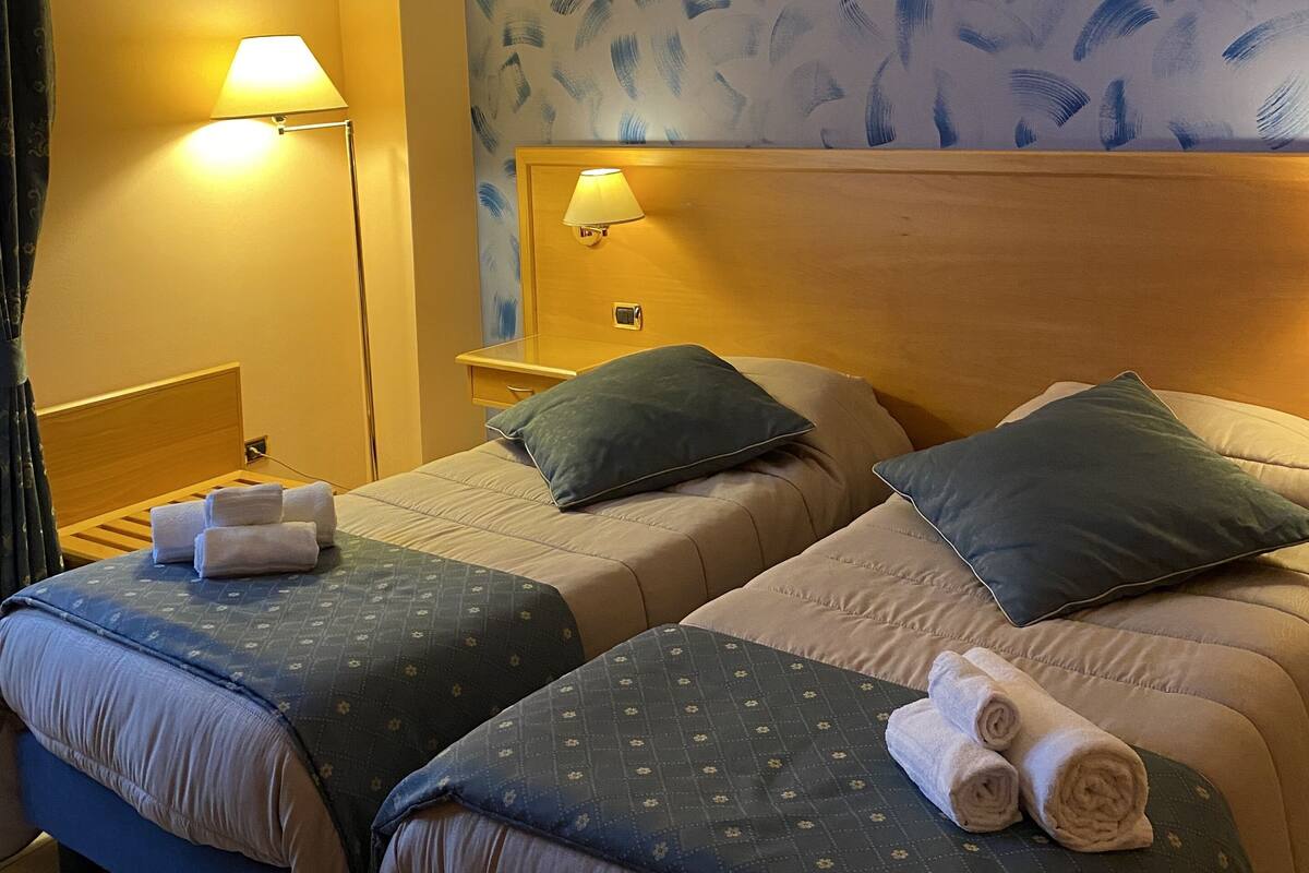 A hotel room with two beds, each with a headboard, bedside lamps, and pillows. There are towels on the beds.