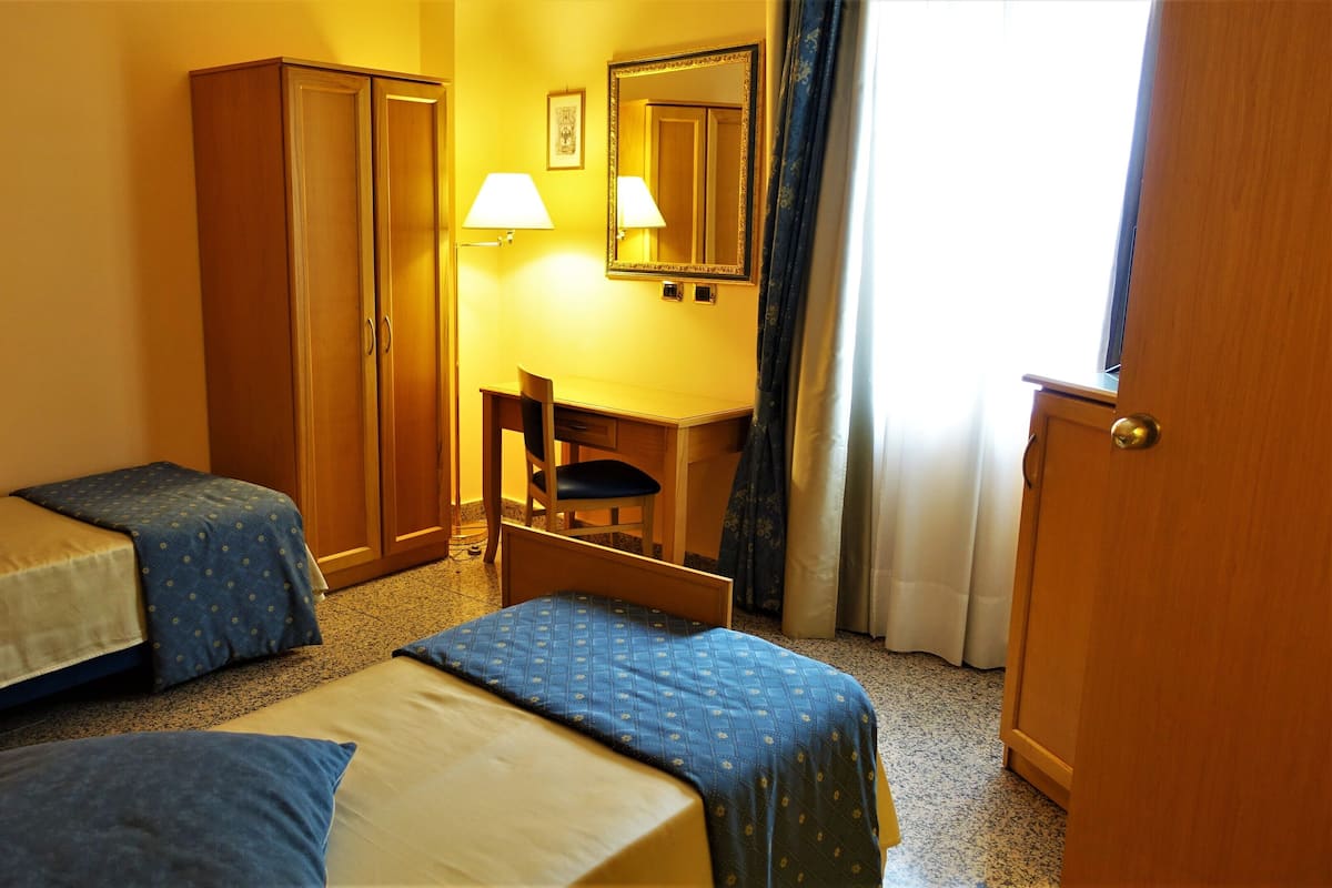 A hotel room with two single beds, a desk with a chair, a wardrobe, a mirror, and a lamp.