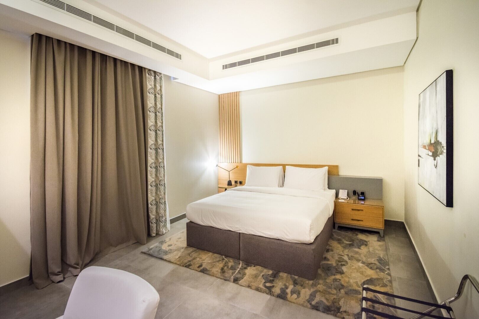 Een hotelkamer met een bed, nachtkastjes, een stoel en een groot raam met gordijnen.