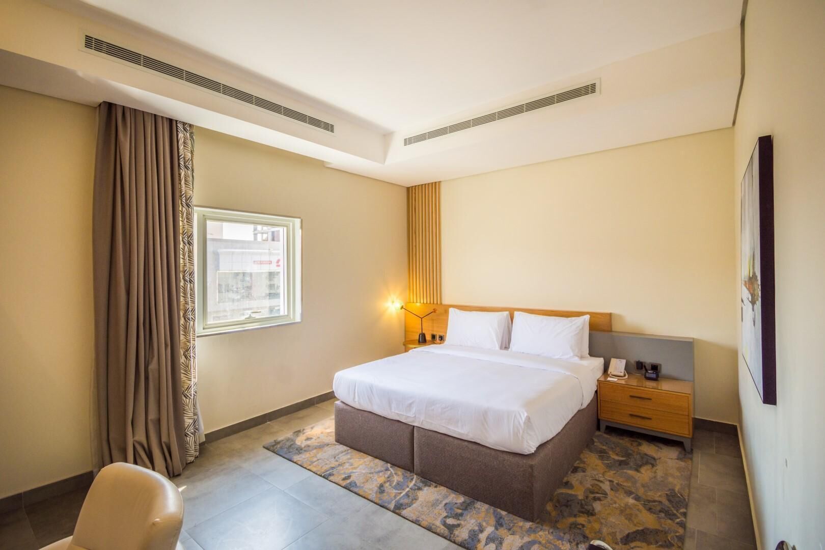 A hotel room with a bed, nightstand, lamp, and a view of the city through the window.