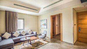A modern living room with a grey sofa, a coffee table, and a grey armchair. There is a door leading to another room and a painting on the wall. - Ewaa Express Hotel - AlRawda (Jeddah)