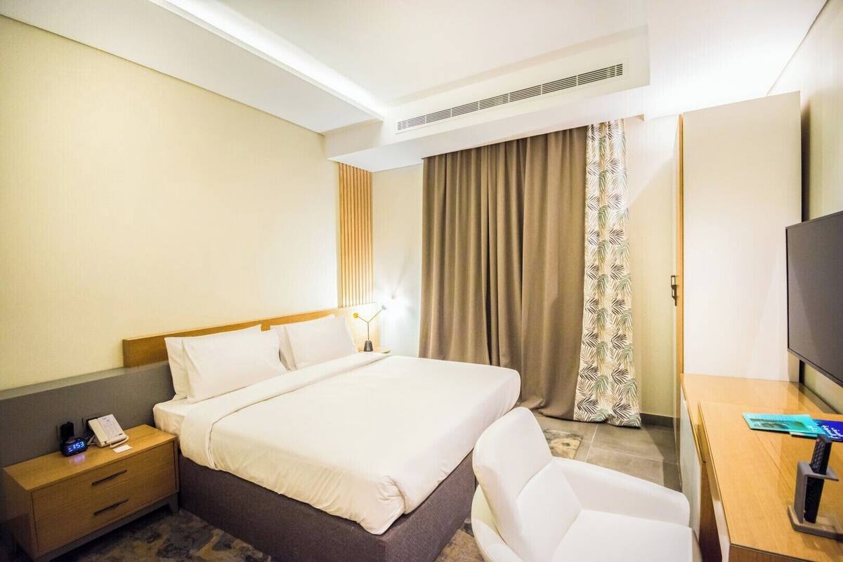 A hotel room with a bed, bedside table, chair, desk, TV, and curtains.
