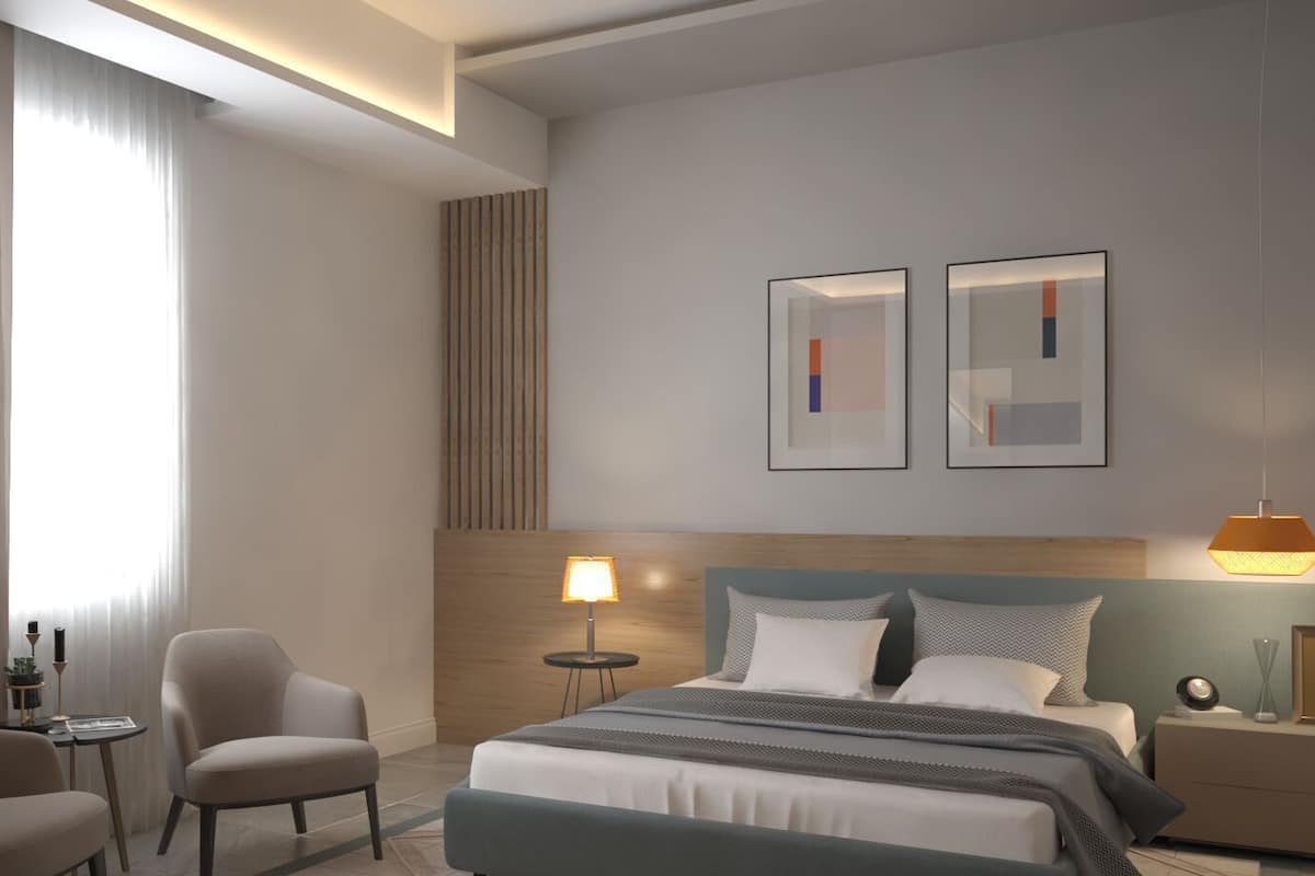 A hotel room with a bed, two chairs, a small table, a lamp, and two framed pictures on the wall.
