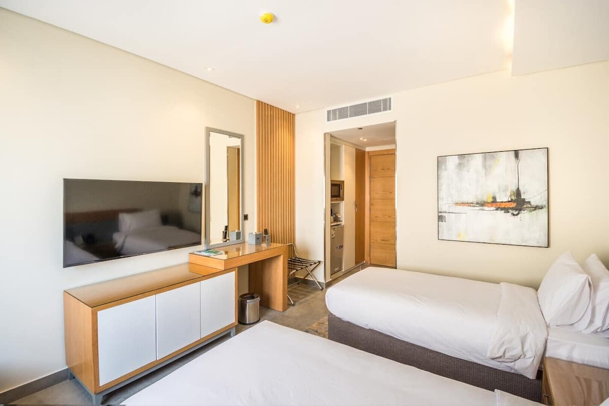 A hotel room with a bed, a desk with a mirror, a TV, a painting, and a wardrobe.
