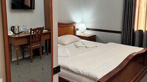 A hotel room with a wooden bed, a desk with a chair, a television, a bedside table with a lamp, and a large mirror. - Stella Hotel (Tashkent)
