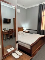 Una habitación de hotel con una cama de madera, un escritorio con silla, un televisor, una mesita de noche con lámpara y un espejo grande.